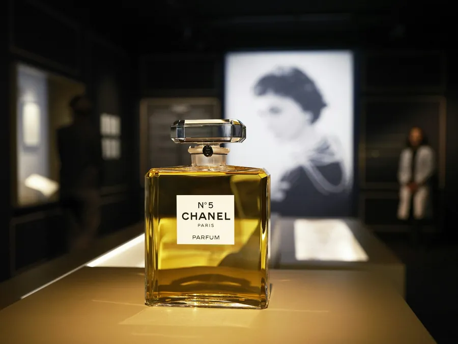 CHANEL N°5 bottle lit on a plinth, with a black and white portrait of Gabrielle Chanel behind it