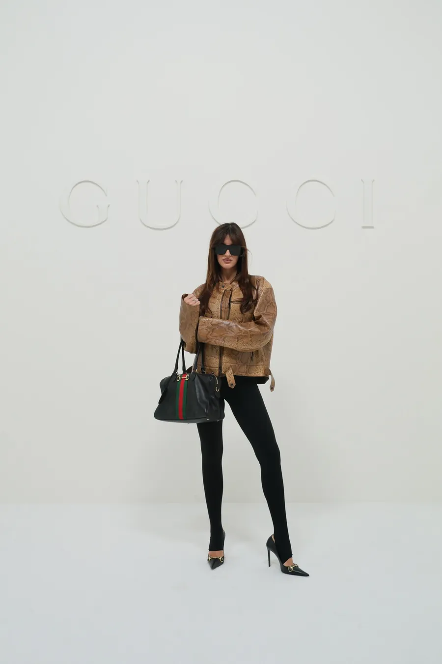 Gucci The Store Show: woman in a camel jacket and black trousers carrying a black Gucci handbag