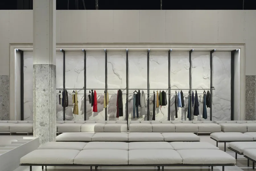 Gucci The Store Show: long clothing rail in front of a white relief wall, with benches and a stone column
